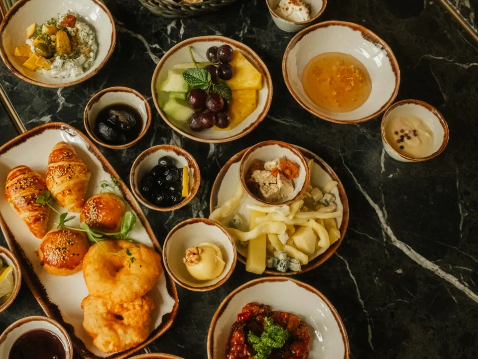 Завтрак на тёмном мраморном столе: круассаны и булочки с кунжутом, пышки, мёд, сыры, оливки, свежие фрукты и варенье в керамических пиалах